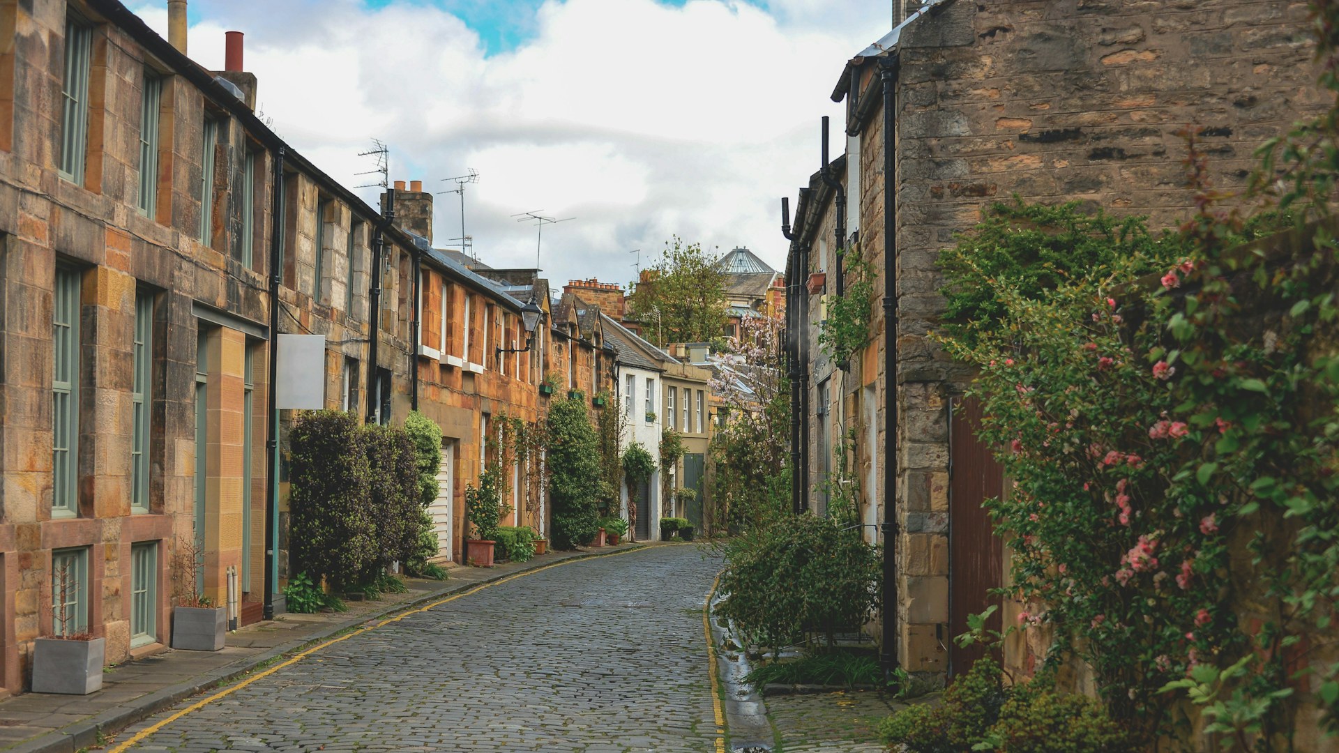 A quiet Edinburgh cobbled lane lined with warm sandstone houses and greenery.