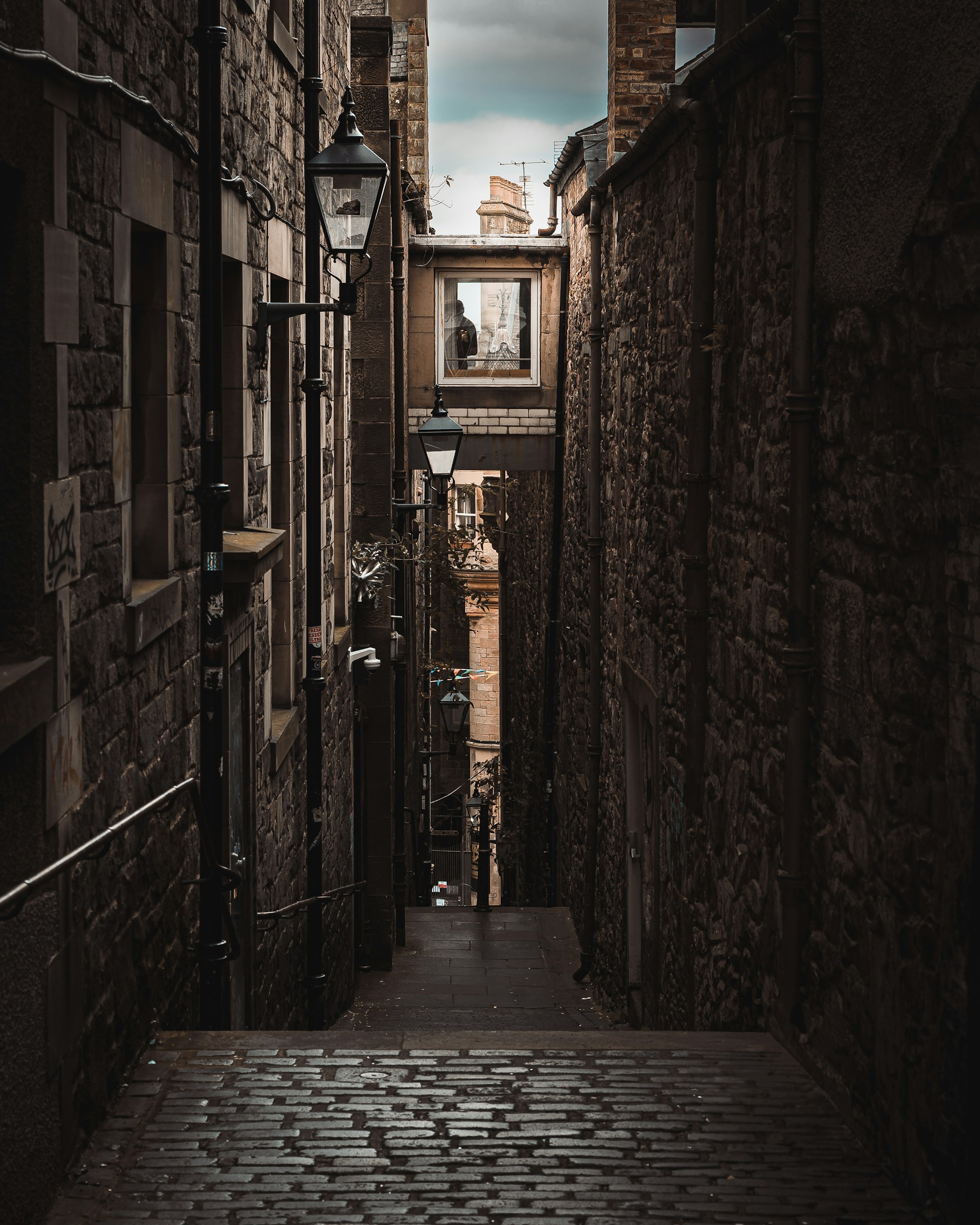 A narrow Edinburgh close of dark stone, steps and ironwork opening onto a brighter street.